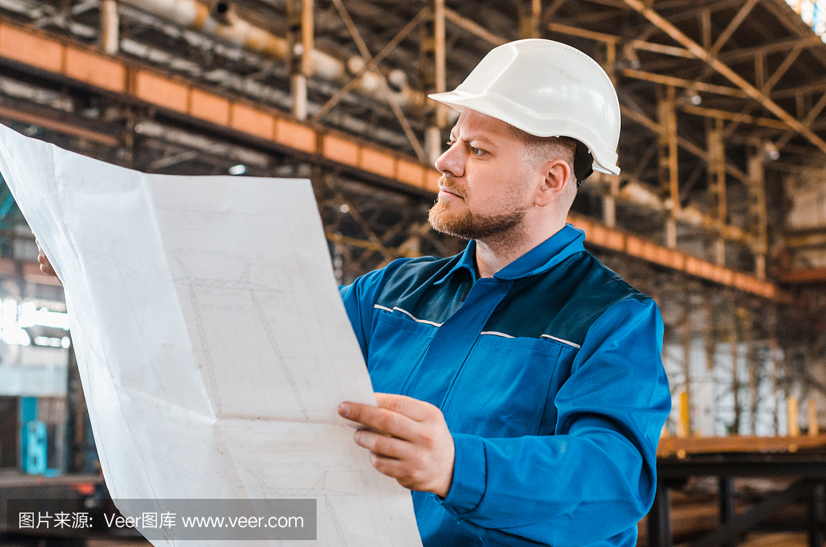 一個穿著工作服、戴著白色頭盔的工人正在檢查一個金屬工廠背景上的圖紙。建筑工人身體很好。男工程師